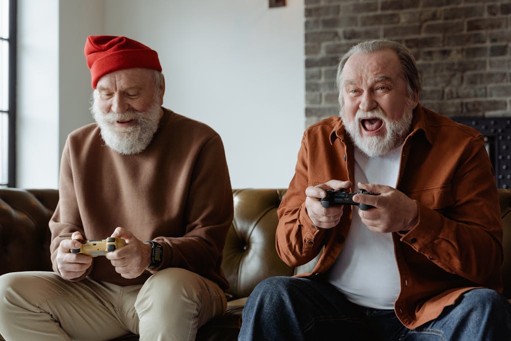 Two senior men laughing and playing video games, sharing joyful moments indoors.